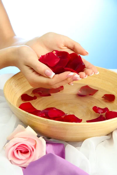 Mani di donna con ciotola di legno di acqua con petali, su sfondo blu — Foto Stock