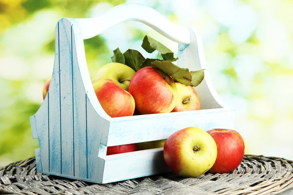 Juicy apples with leaves in wooden basket, on green background — Stock Photo, Image