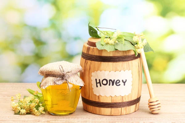 Jar and barrel with linden honey and flowers on wooden table on green background — Stock Photo, Image