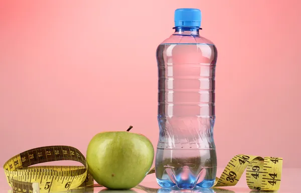 Garrafa de água, maçã e fita métrica em fundo rosa — Fotografia de Stock