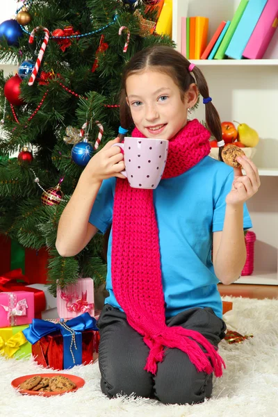 Niña con bufanda rosa y taza de bebida caliente sentada cerca del árbol de Navidad —  Fotos de Stock