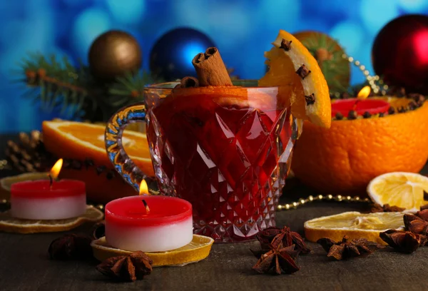 Fragrant mulled wine in glass with spices and oranges around on wooden table on blue background — Stock Photo, Image