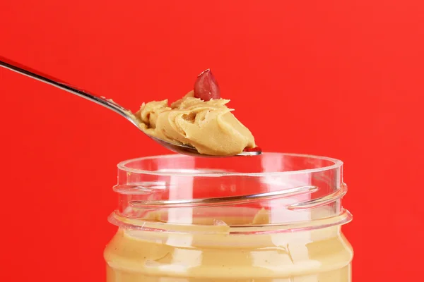Deliciosa manteiga de amendoim em frasco e colher no fundo vermelho — Fotografia de Stock