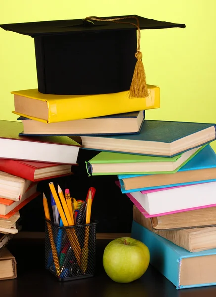 Libros y gorra magister contra pizarra escolar sobre mesa de madera sobre fondo verde — Foto de Stock