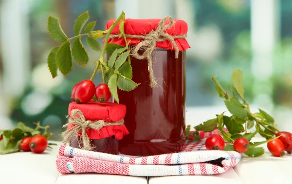 Potten met hip rozen jam en rijpe bessen, op houten tafel — Stockfoto