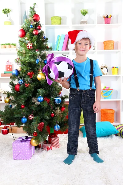Santa kalap kisfiú áll, közel a futball-labdát karácsonyi fa — Stock Fotó