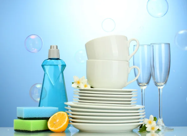Platos limpios vacíos, vasos y tazas con líquido lavavajillas, esponjas y limón sobre fondo azul — Foto de Stock