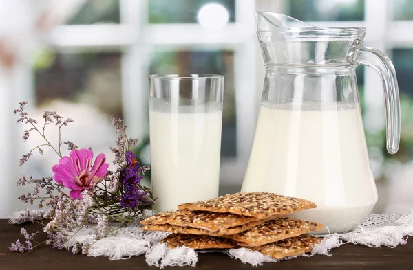 Кувшин и стакан молока с печеньем на деревянном столе на фоне окна — стоковое фото
