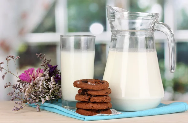 Džbán a sklenice mléka s cookies na dřevěný stůl na pozadí okna — Stock fotografie