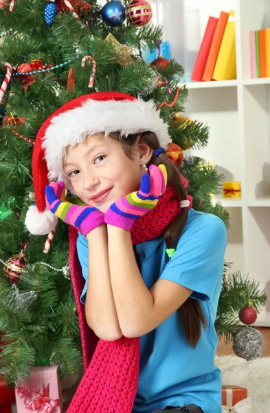 Niña con bufanda rosa y guantes multicolores sentada cerca del árbol de Navidad — Foto de Stock