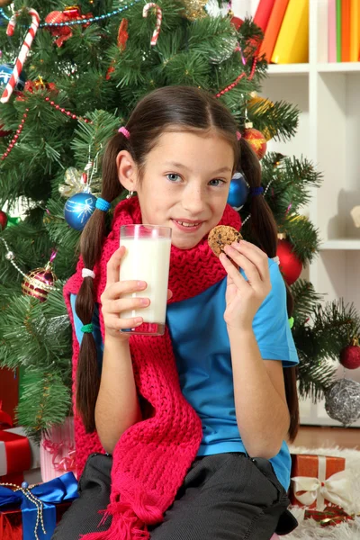 Kis lány rózsaszín sál és pohár tejet karácsonyfa közelében ült — Stock Fotó