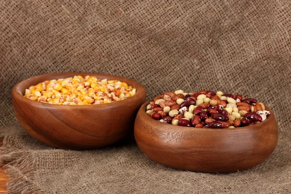 Nezpracovaná kukuřice a fazole v dřevěných misek na tabulce na pozadí pytloviny — Stock fotografie
