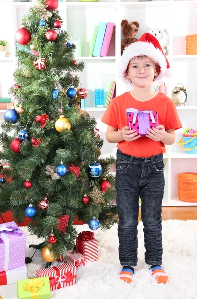 Kleine jongen in Kerstman hoed staat in de buurt van kerstboom met cadeau in zijn handen — Stockfoto