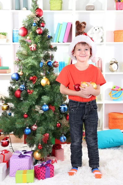 Little boy in Santa hat decorates Christmas tree in room Stock Image