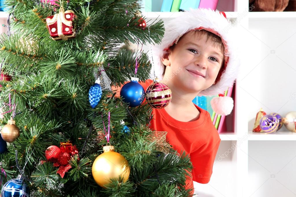Little boy in Santa hat peeks out from behind Christmas tree
