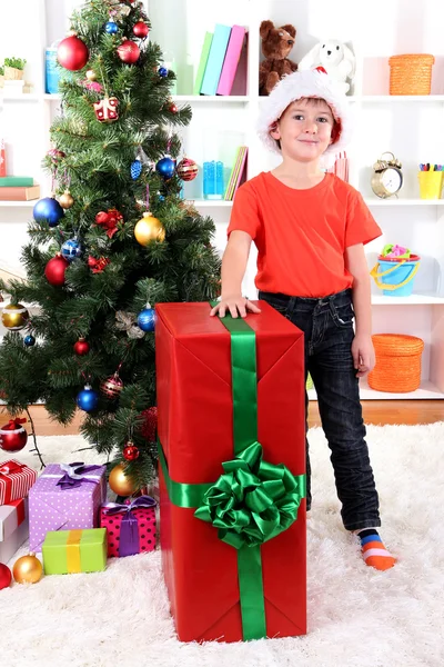 Child in Santa hat near Christmas tree with big gift Stock Photo