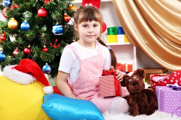 Little girl sits near a Christmas tree with gift in hand in festively decorated room — Stock Photo, Image