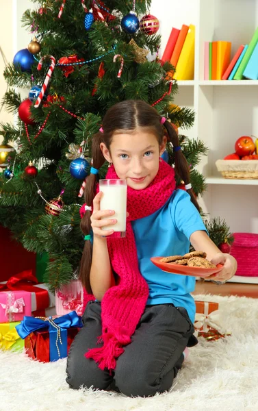 Liten flicka med rosa halsduk och glas mjölk sitter nära julgran — Stockfoto