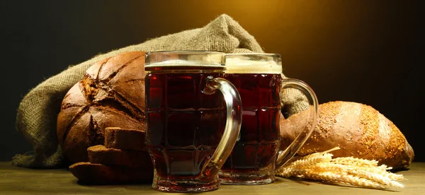 Tankards of kvass and rye breads with ears, on wooden table on brown background — Stock Photo, Image
