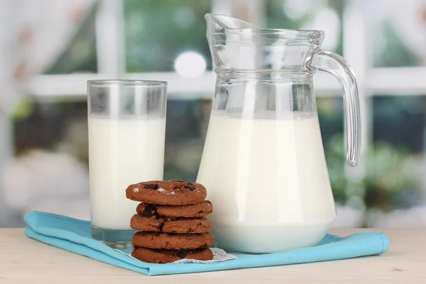 Džbán a sklenice mléka s cookies na dřevěný stůl na pozadí okna — Stock fotografie