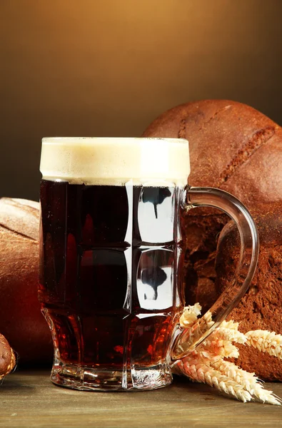 Tankard of kvass and rye breads with ears, on wooden table on brown background — Stock Photo, Image