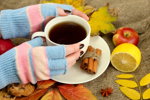 Hands holding cup of hot drink and autumn leaves, on burlap background — Stock Photo, Image