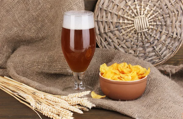 Стакан пива с тарелкой чипсов на деревянном столе на фоне мешка — стоковое фото