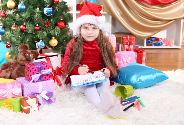 Beautiful little girl in red dress writes letter to Santa Claus in festively decorated room — Stock Photo, Image