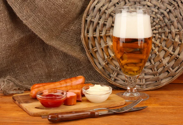 Cerveja e salsichas grelhadas em mesa de madeira sobre fundo de pano de saco — Fotografia de Stock