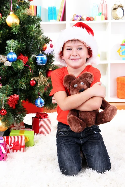 Niño en el sombrero de Santa se sienta cerca del árbol de Navidad con regalo en las manos — Foto de Stock