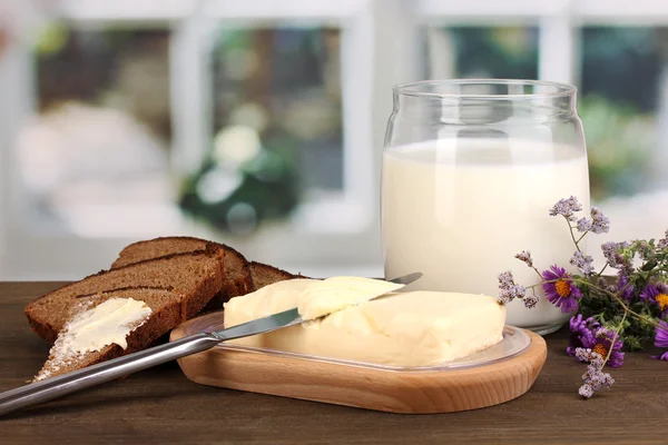 Burro su supporto di legno circondato da pane e latte su tavolo di legno su sfondo finestra — Foto Stock