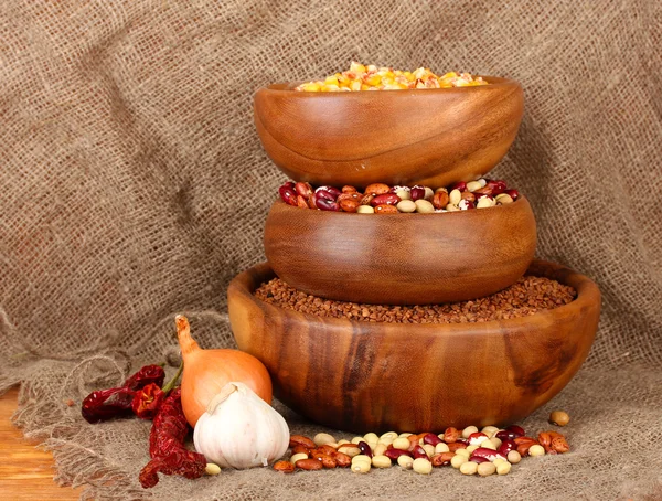 Nezpracovaná kukuřice, pohanka a fazole v dřevěných misek na tabulce na pozadí pytloviny — Stock fotografie