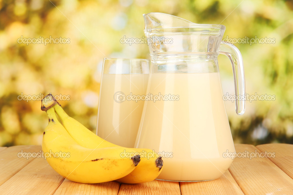 Full glass and jug of banana juice and bananas on wooden table outdoor