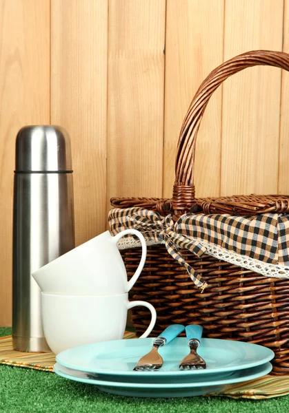 Metal thermos with cups, plates and basket on grass on wooden background — Stock Photo, Image