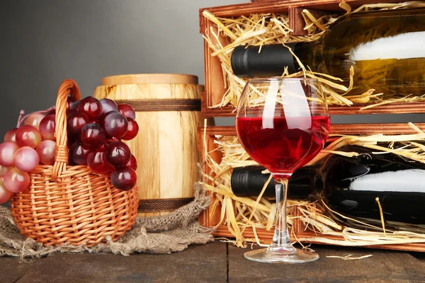 Cassa in legno con bottiglie di vino, botte, bicchiere da vino e uva su tavolo in legno su sfondo grigio — Foto Stock