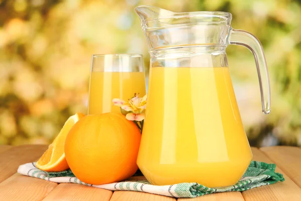 Copo completo e jarro de suco de laranja e laranjas na mesa de madeira ao ar livre — Fotografia de Stock
