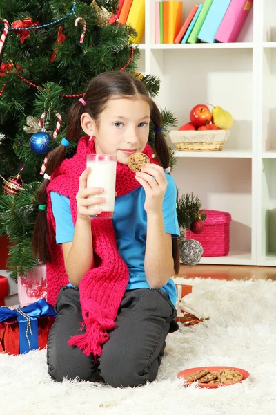 Kis lány rózsaszín sál és pohár tejet karácsonyfa közelében ült — Stock Fotó