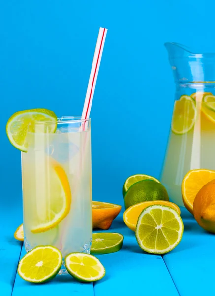 Limonada cítrica em jarro e vidro de citrinos em torno de mesa de madeira sobre fundo azul — Fotografia de Stock