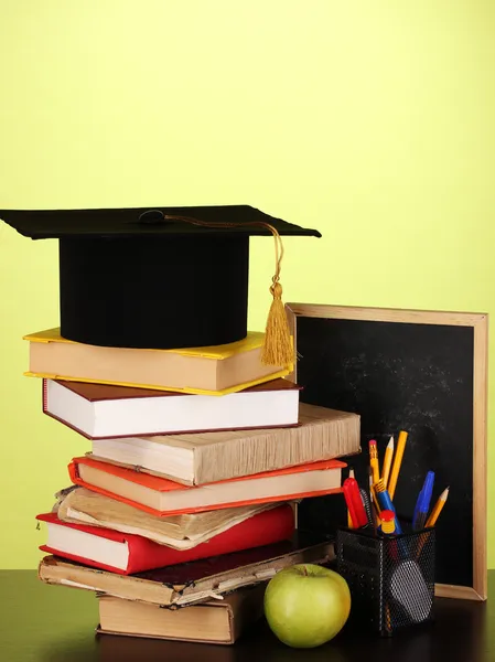 Books and magister cap against school board on wooden table on green background — Stock Photo, Image