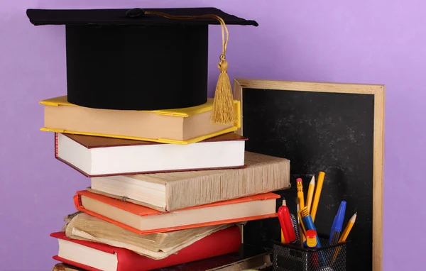 Books and magister cap against school board on wooden table on purple background — Stock Photo, Image