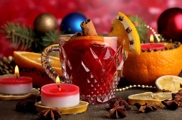 Fragrant mulled wine in glass with spices and oranges around on wooden table on red background — Stock Photo, Image