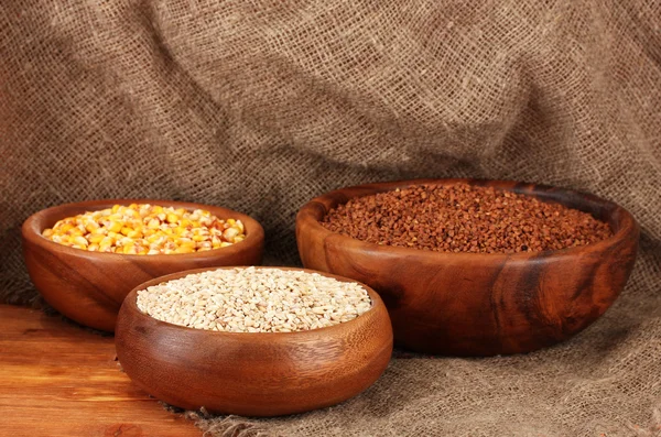 Maïs cru, sarrasin et blé dans des bols en bois sur la table sur fond de sac — Photo
