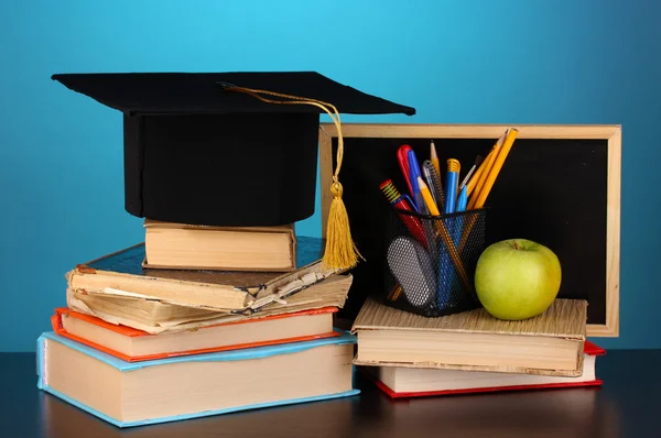 Books and magister cap against school board on wooden table on blue background — Stock Photo, Image