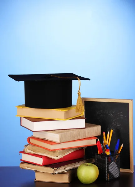 Books and magister cap against school board on wooden table on blue background — Stock Photo, Image