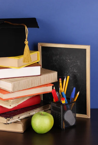 Books and magister cap against school board on wooden table on blue background — Stock Photo, Image