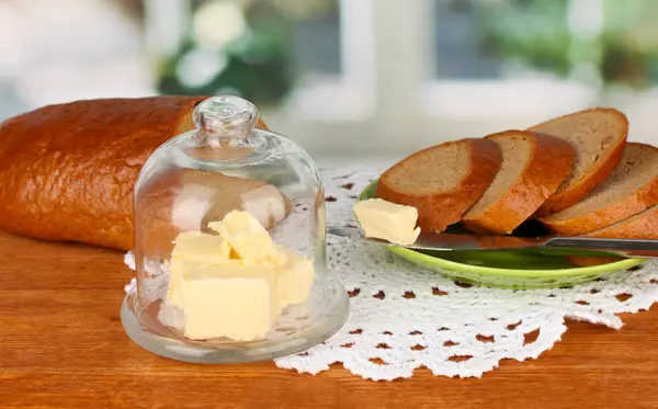 Mantequilla en platillo de vidrio con tapa de vidrio rodeada de pan, sobre fondo brillante — Foto de Stock