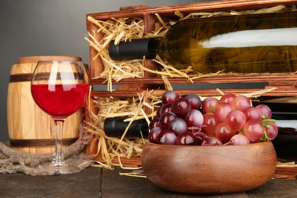 Cassa in legno con bottiglie di vino, botte, bicchiere da vino e uva su tavolo in legno su sfondo grigio — Foto Stock