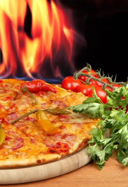 Smakelijke pepperoni pizza met groenten op houten bord op vlam achtergrond — Stockfoto