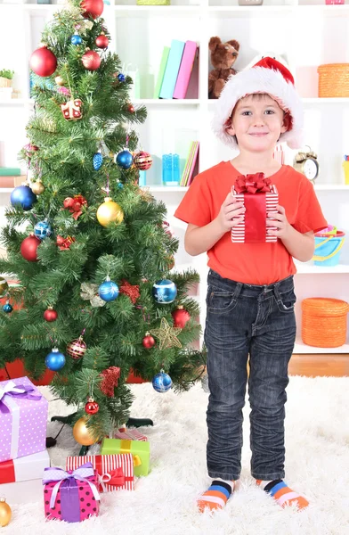 Kleine jongen in Kerstman hoed staat in de buurt van kerstboom met cadeau in zijn handen — Stockfoto