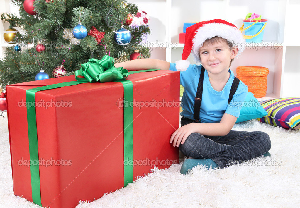 Child in Santa hat sits near Christmas tree with big gift in hands